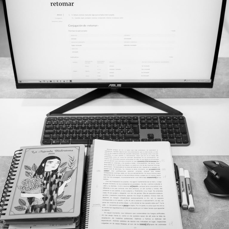 Mesa con un ordenador, un libro abierto y material de escritura.